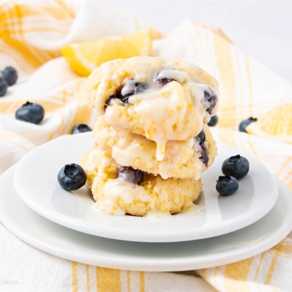 A stack of three glazed blueberry lemon cookies sits on a white plate, surrounded by fresh blueberries, with a yellow and white checkered cloth and a lemon wedge in the background.