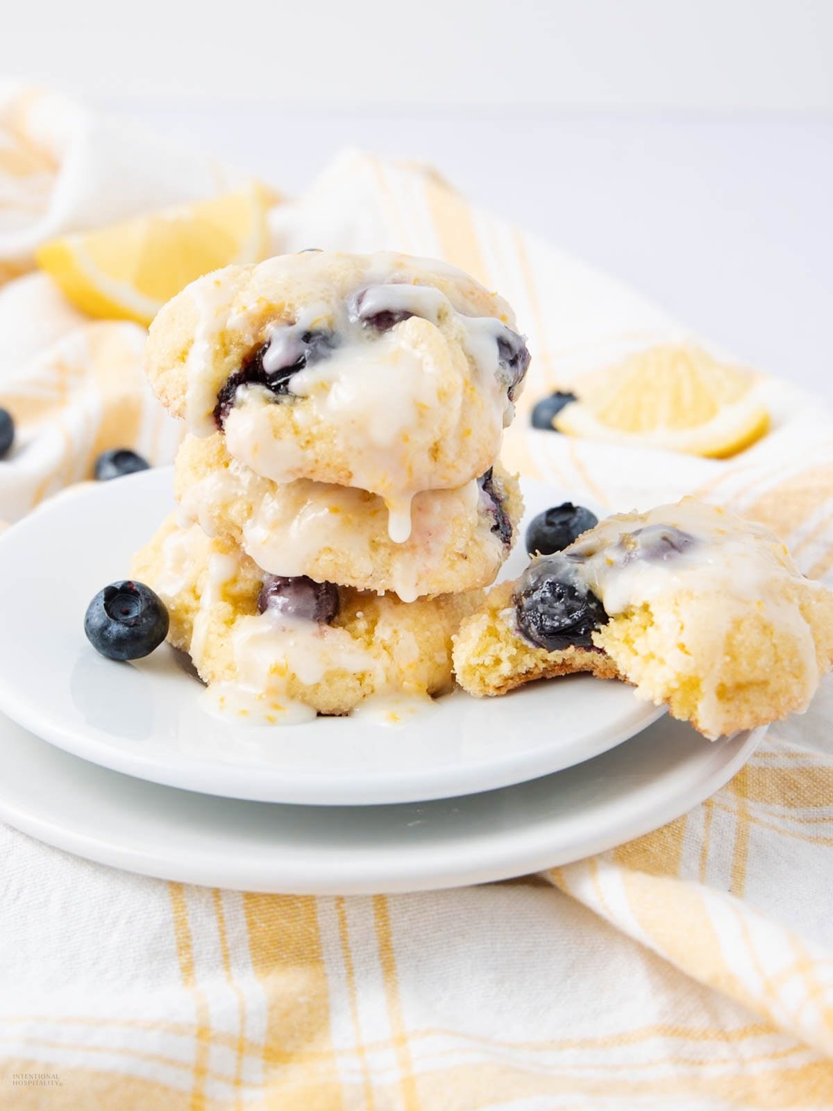 A stack of three glazed blueberry cookies sits on a white plate, with fresh blueberries and a lemon wedge nearby. One cookie is broken open, revealing the blueberries inside. A yellow plaid cloth is in the background.