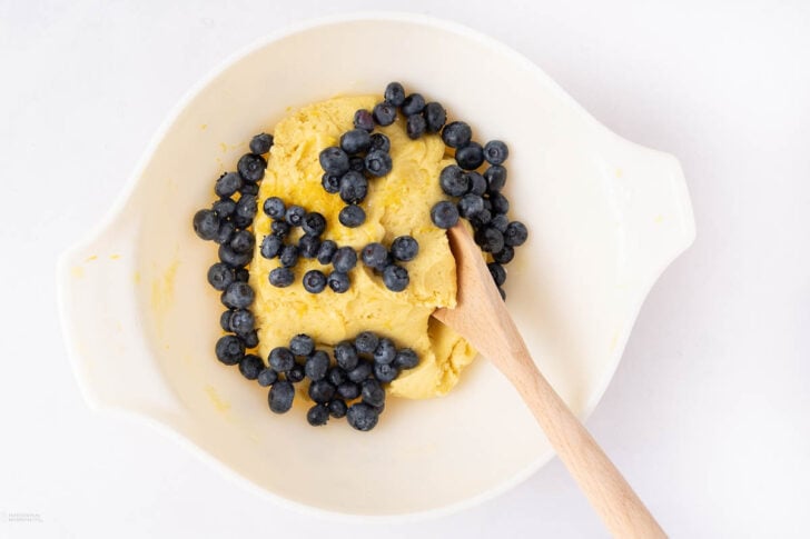 A white mixing bowl containing yellow cookie dough and fresh blueberries, with a wooden spoon resting inside the bowl.