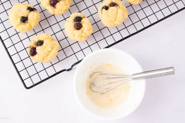 A cooling rack with several mini blueberry muffins sits next to a white bowl containing glaze and a small whisk on a white surface.
