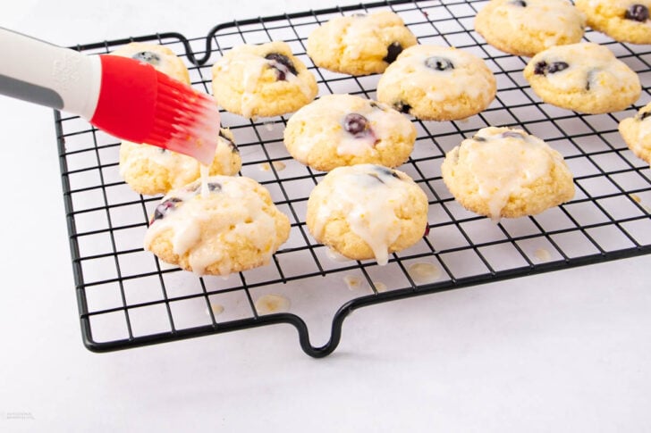 A hand uses a red silicone brush to spread glaze over freshly baked blueberry cookies cooling on a black wire rack. Some cookies are already covered with glaze, and a few drips are visible on the white surface below.