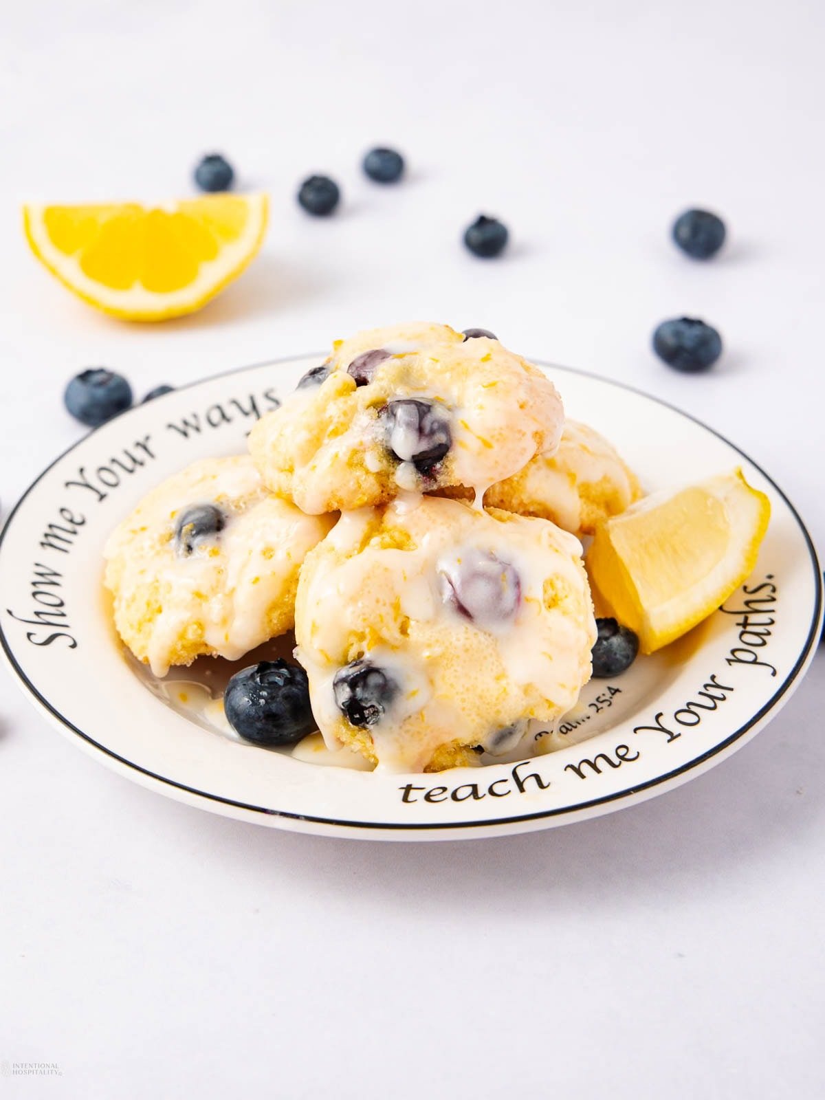 A plate of blueberry lemon cookies is stacked with fresh blueberries and lemon wedges, surrounded by scattered blueberries and a lemon slice. The plate has the text “Show me Your ways… teach me Your paths.”.