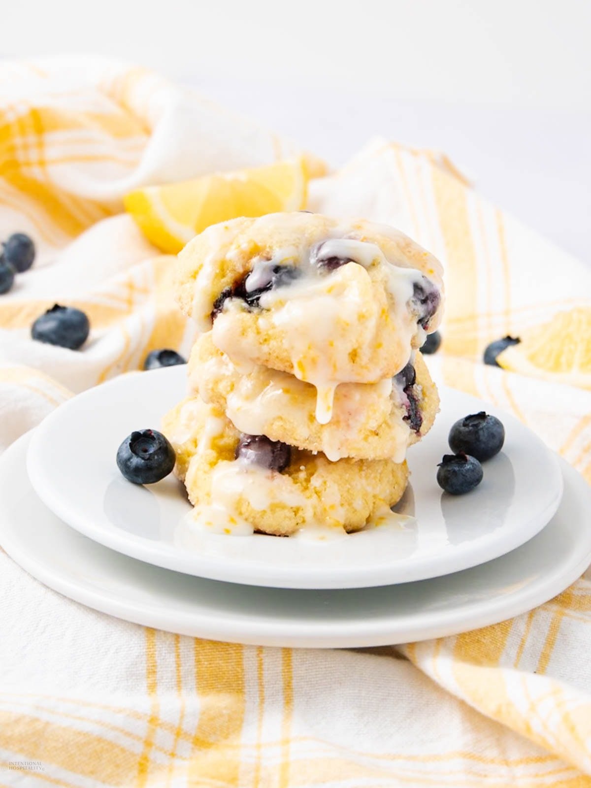 A stack of three glazed blueberry lemon cookies sits on a white plate, with fresh blueberries scattered around and a yellow plaid cloth in the background.