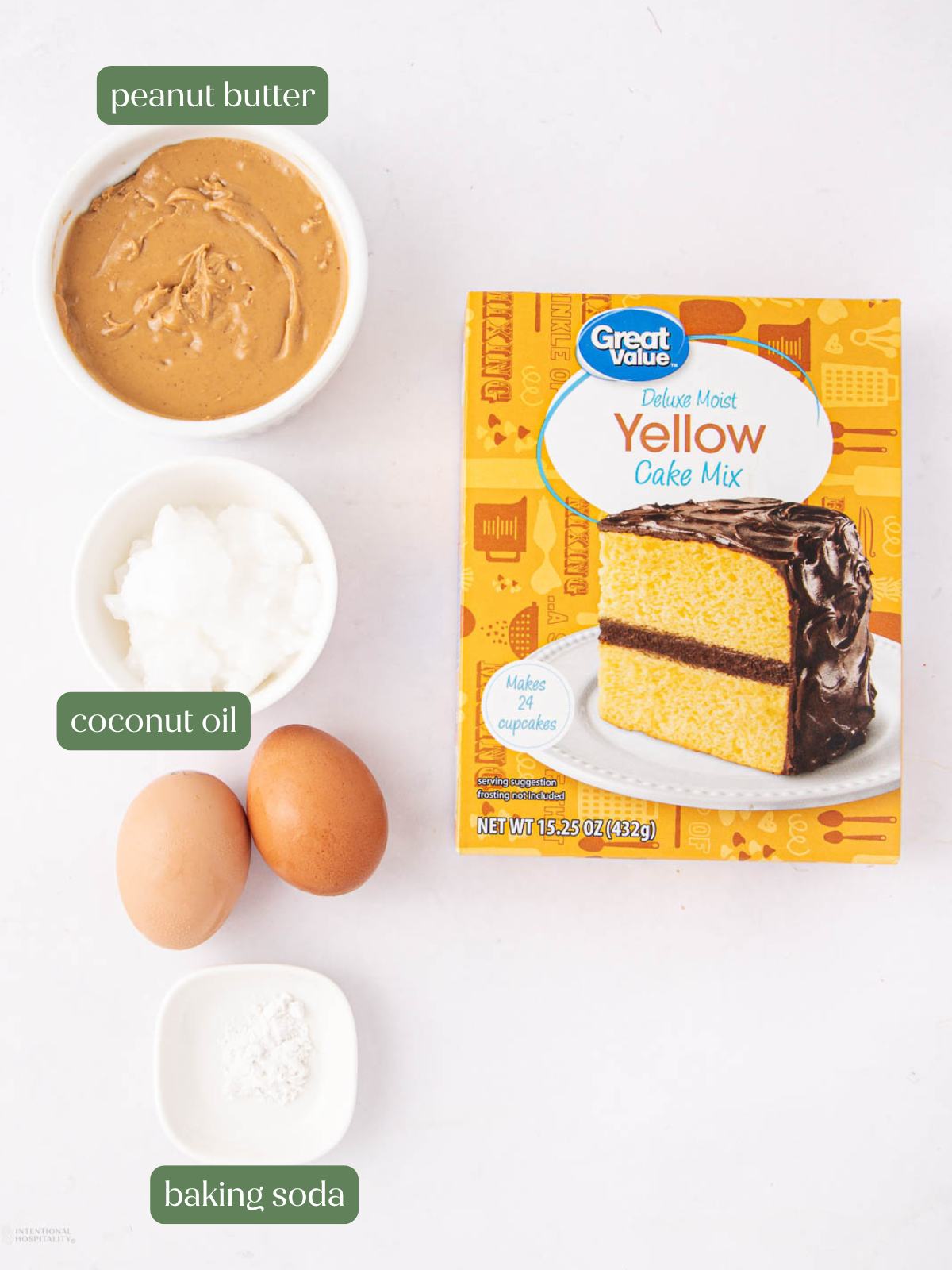 A box of yellow cake mix, a bowl of peanut butter, a bowl of coconut oil, two eggs, and a small dish of baking soda are arranged on a white surface. Each ingredient is labeled.