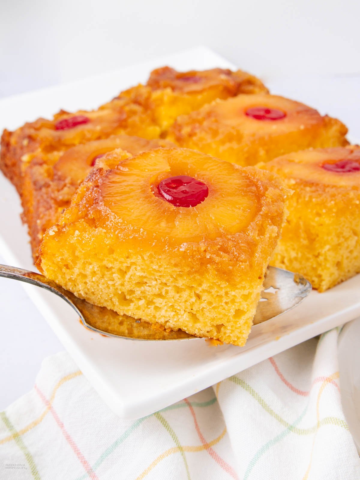 A slice of pineapple upside-down cake topped with a pineapple ring and cherry is being lifted from a white plate holding several more slices. A colorful, checkered cloth sits underneath the plate.