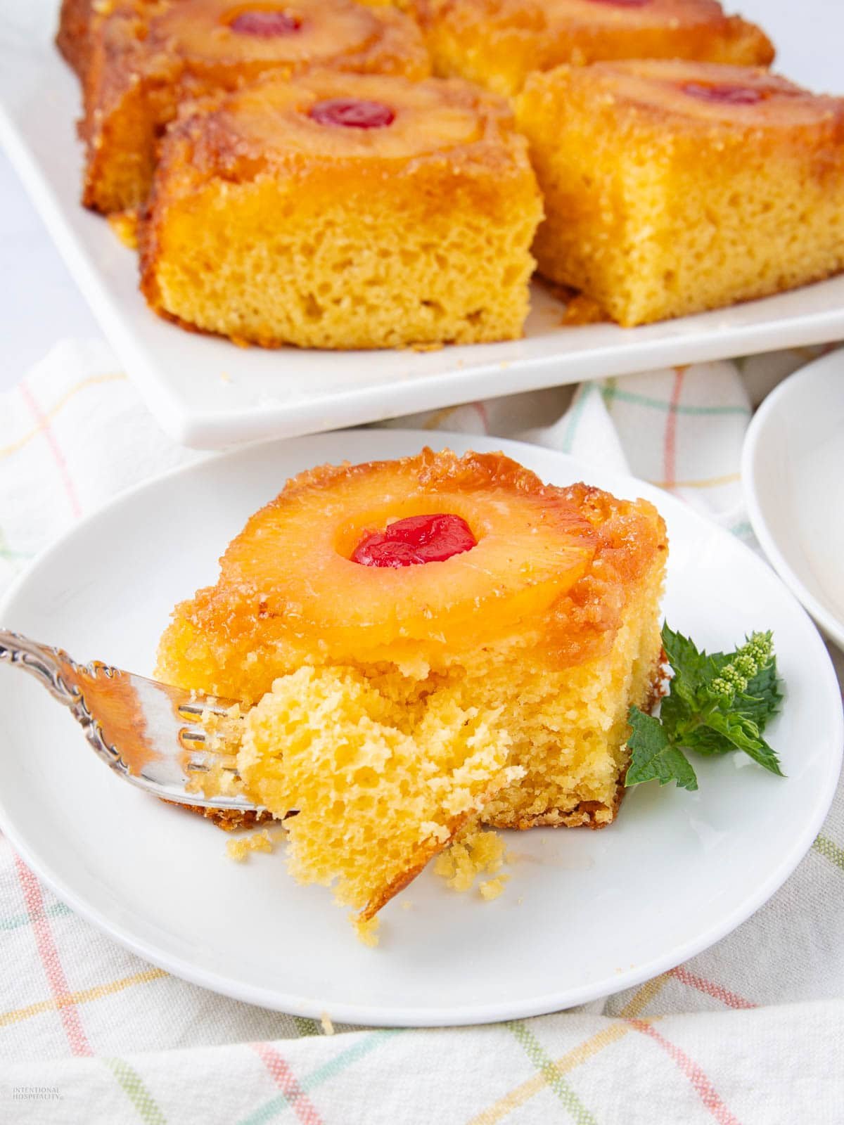 A slice of pineapple upside-down cake with a cherry on top sits on a white plate, with a fork cutting a bite. More cake slices are visible in the background on a tray. A mint garnish is on the plate.