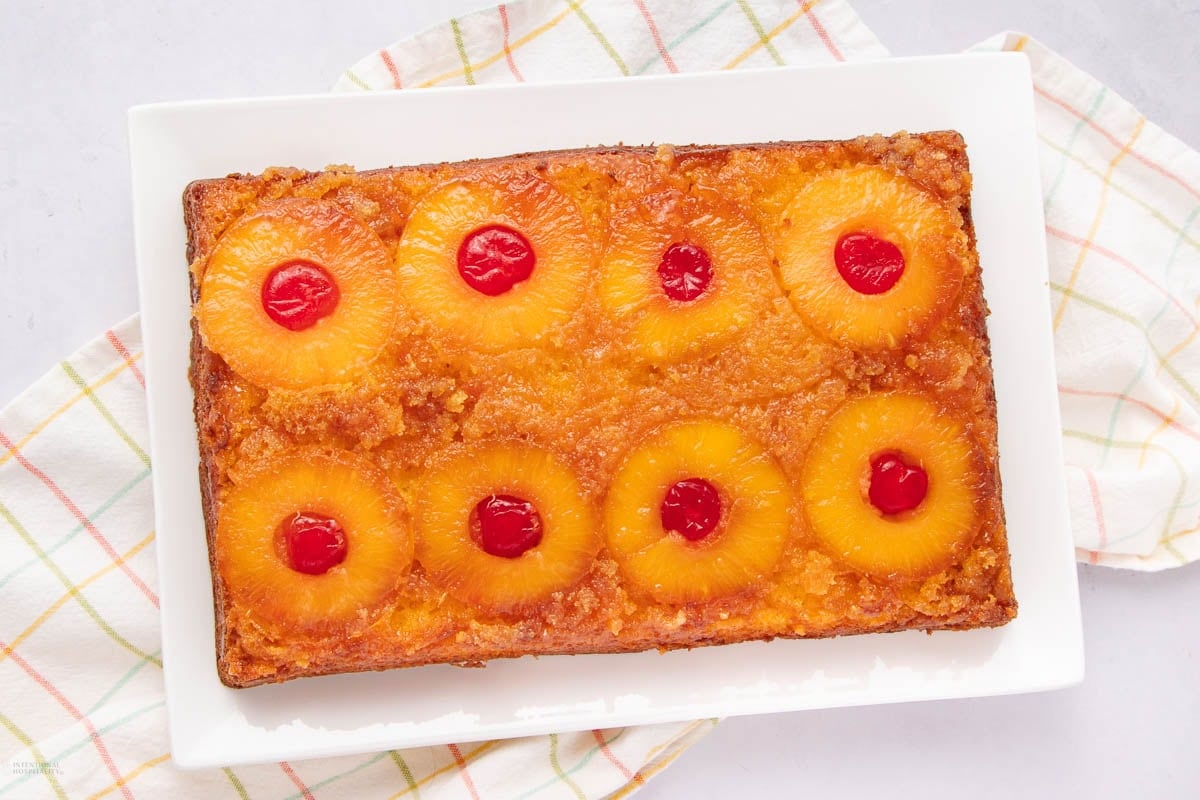 A rectangular pineapple upside-down cake topped with pineapple rings and maraschino cherries sits on a white plate over a plaid kitchen towel.
