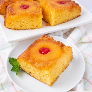 A slice of pineapple upside-down cake topped with a pineapple ring and cherry, served on a white plate with a mint garnish. More cake slices are on a white tray in the background.
