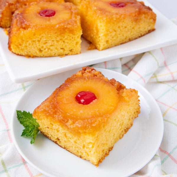 A slice of pineapple upside-down cake topped with a pineapple ring and cherry, served on a white plate with a mint garnish. More cake slices are on a white tray in the background.