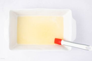 A white rectangular baking dish with a melted butter coating, being spread with a red and white pastry brush, on a white surface.