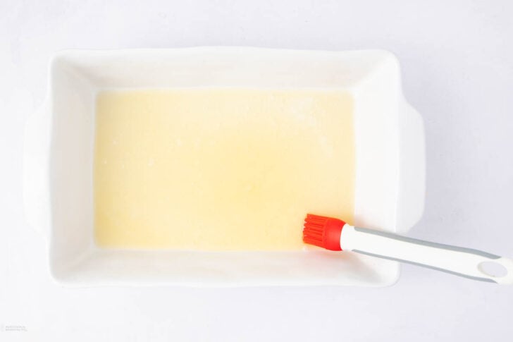 A white rectangular baking dish with a melted butter coating, being spread with a red and white pastry brush, on a white surface.