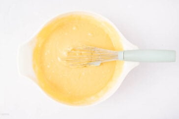 A white mixing bowl filled with yellow cake batter, with a metal whisk resting inside, placed on a white surface.