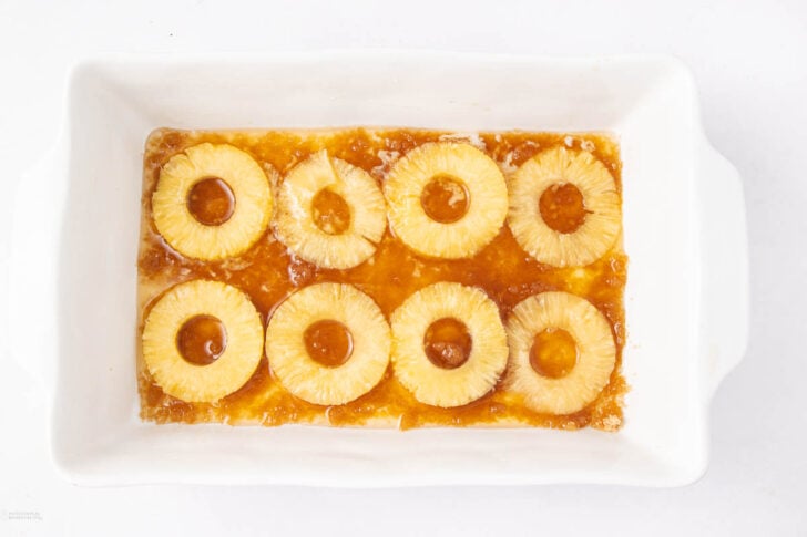 A white rectangular baking dish containing a layer of pineapple rings arranged over a caramelized brown sugar mixture.