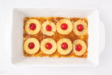 A rectangular white baking dish containing eight pineapple rings, each topped with a red maraschino cherry, arranged in rows on a layer of brown sugar mixture.