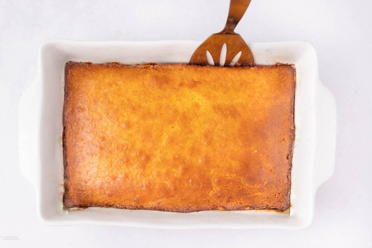 A rectangular baked casserole with a golden-brown top sits in a white dish. A slotted serving spatula is placed under one corner, ready to serve. The background is a clean, light surface.