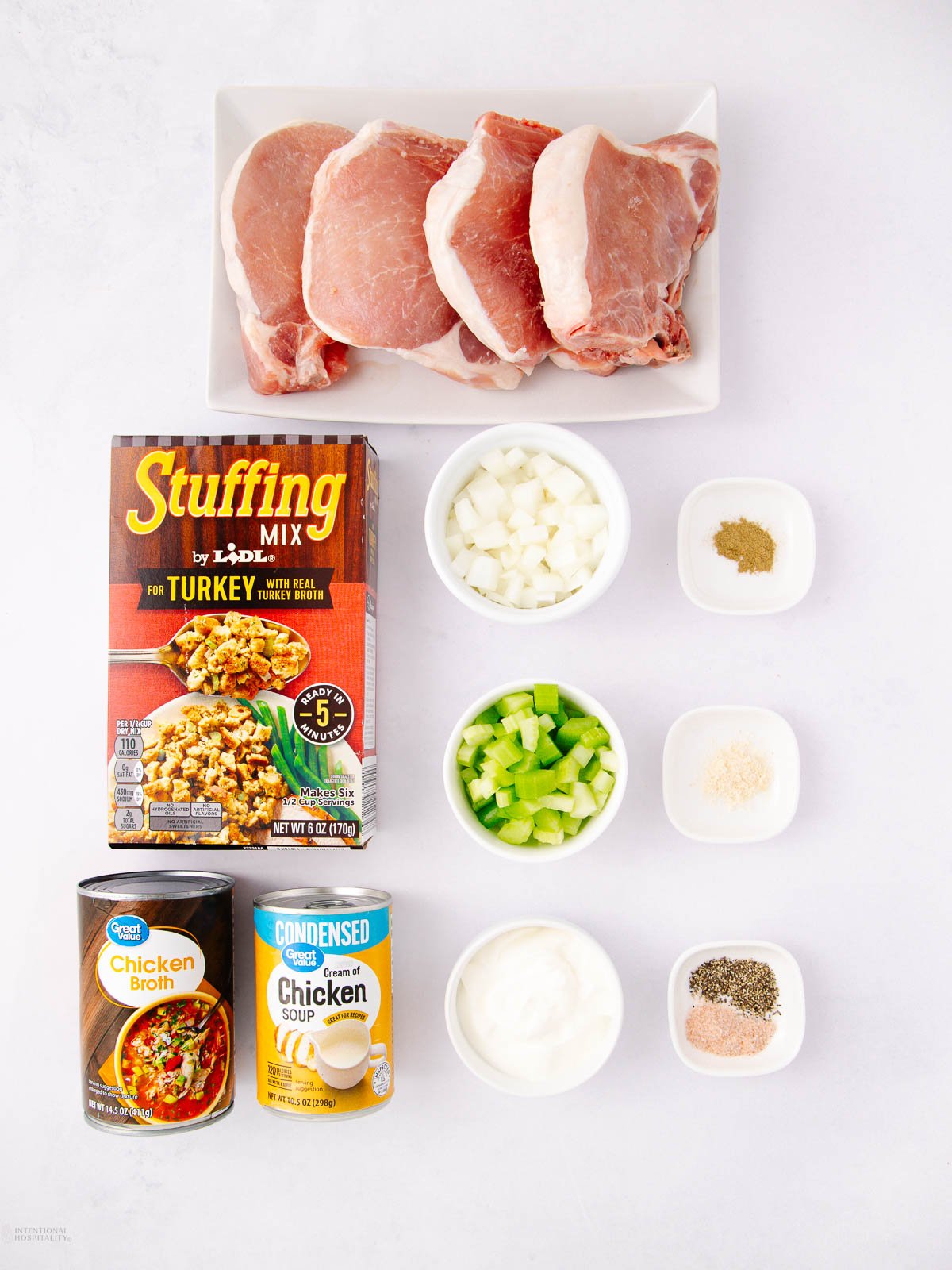 Overhead view of pork chops, turkey stuffing mix, diced onions, diced celery, chicken broth, cream of chicken soup, sour cream, and small bowls of spices arranged on a white surface.