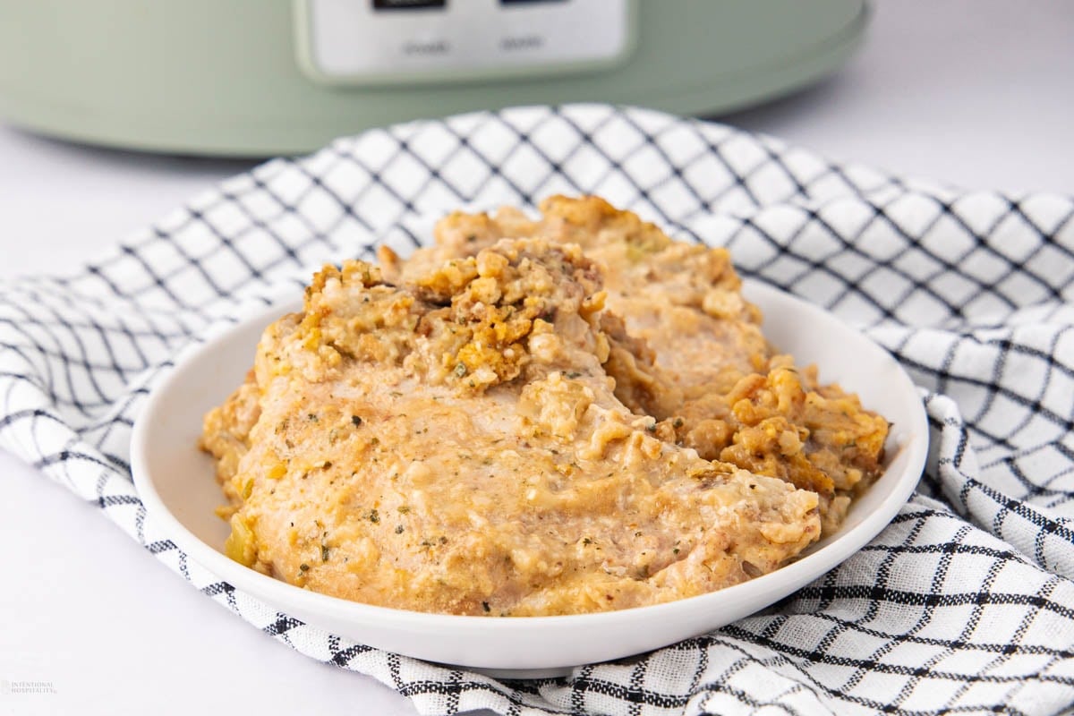 A white plate holds two pieces of creamy, seasoned chicken and rice casserole, sitting on a black-and-white checkered cloth with part of a slow cooker visible in the background.