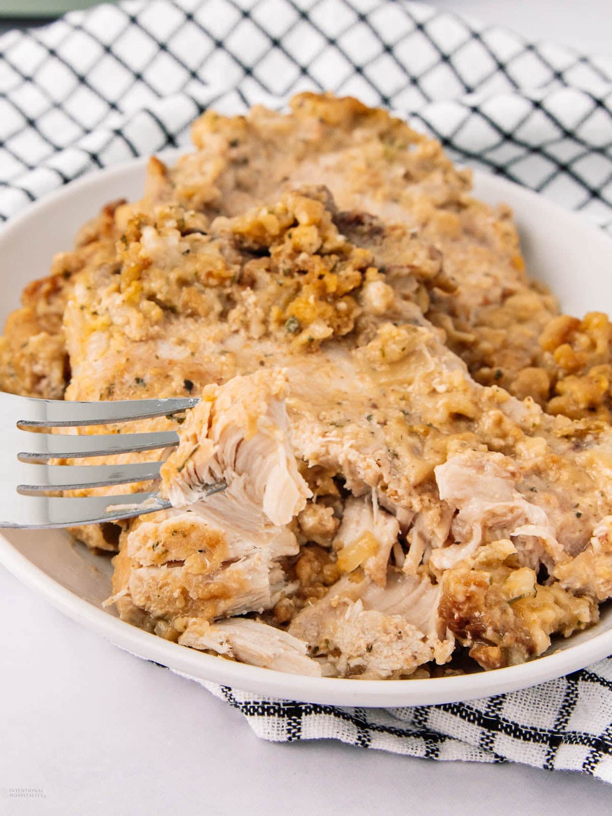 A fork cutting into a serving of creamy chicken and stuffing casserole on a white plate, with a black and white checkered napkin underneath. The dish looks moist and seasoned, with visible chunks of chicken and stuffing.