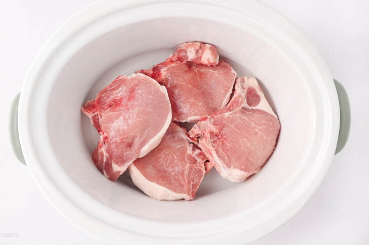 Four raw pork chops placed inside a white slow cooker on a white surface, ready to be cooked.