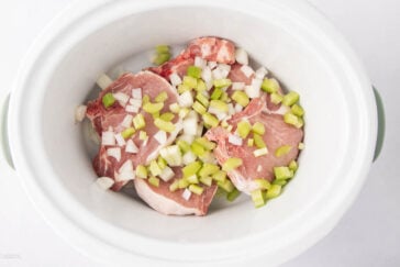 Raw pork chops topped with chopped onions and celery pieces are placed inside a white slow cooker on a white background.
