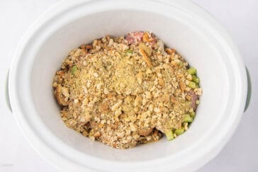 A white slow cooker filled with raw ingredients, including chopped green bell peppers, chicken pieces, and a layer of crushed crackers and seasonings on top.
