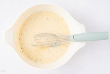 A white mixing bowl filled with a light-colored, speckled batter or sauce, with a whisk featuring a light blue handle resting inside.