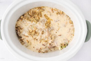 A slow cooker filled with a creamy mixture of sauce, crumbled stuffing, and visible pieces of meat or vegetables, ready to be cooked.