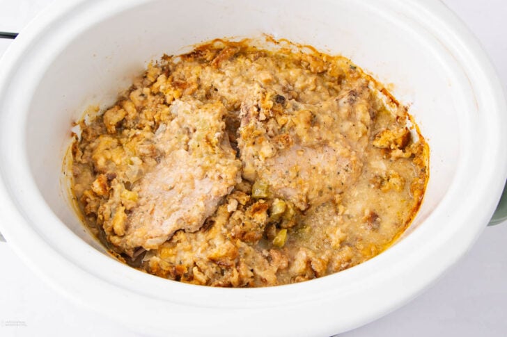 A close-up of a slow cooker filled with cooked pork chops and stuffing, showing tender meat surrounded by a moist, crumbly bread stuffing mixture with visible spices and herbs.