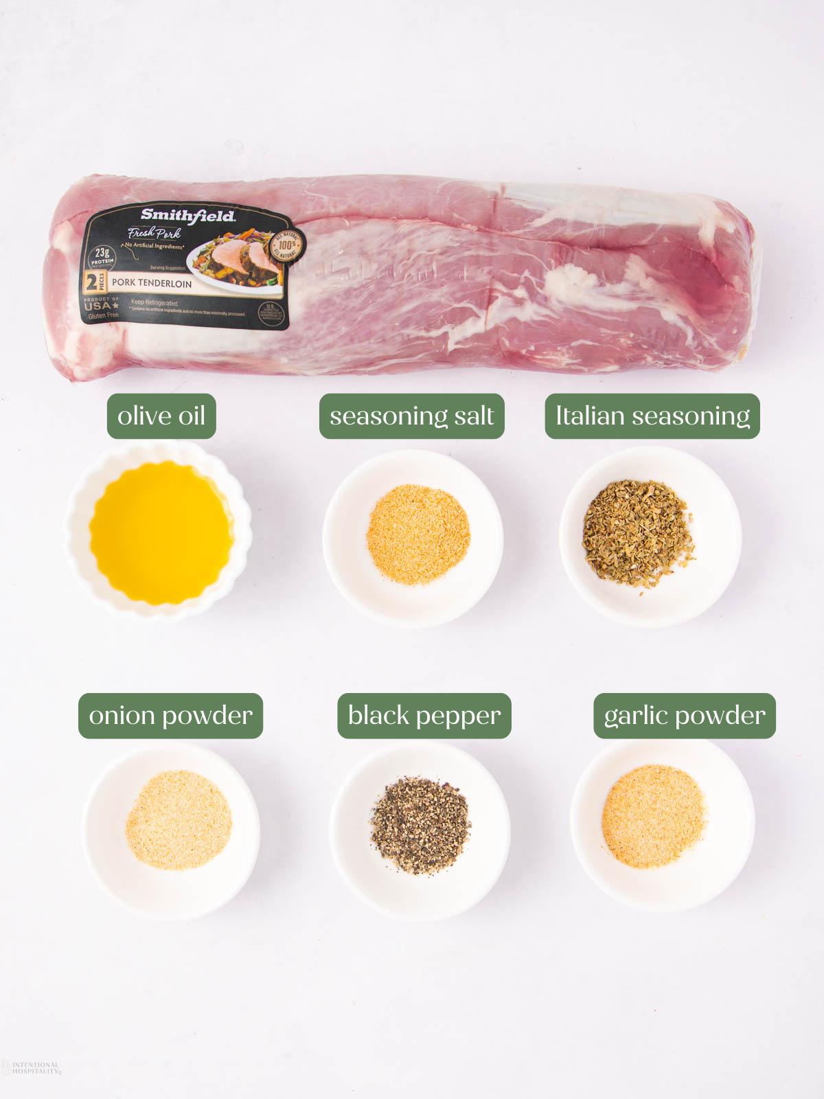 Raw pork tenderloin and six white bowls containing olive oil, seasoning salt, Italian seasoning, onion powder, black pepper, and garlic powder, each labeled accordingly, arranged on a white surface.