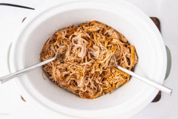 Shredded cooked meat in a white slow cooker with two metal forks, ready for serving. The meat appears seasoned and tender.