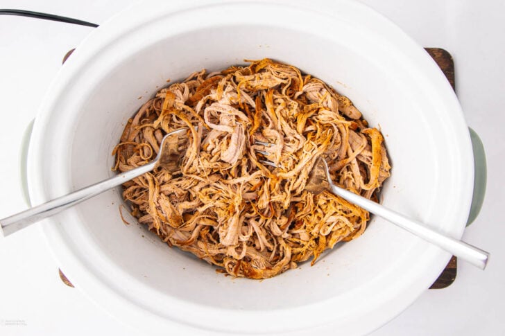 Shredded cooked meat in a white slow cooker with two metal forks, ready for serving. The meat appears seasoned and tender.