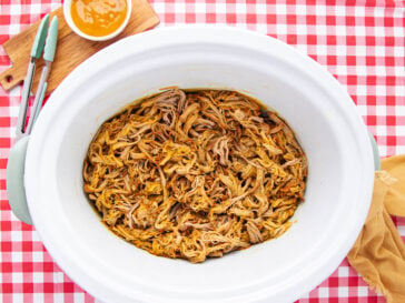 Shredded barbecue pulled pork in a slow cooker on a red and white checkered tablecloth, with tongs, a yellow cloth, and a bowl of sauce nearby.