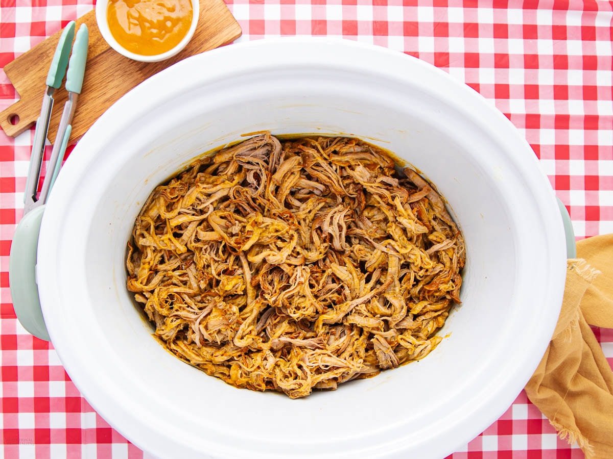 Shredded barbecue pulled pork in a slow cooker on a red and white checkered tablecloth, with tongs, a yellow cloth, and a bowl of sauce nearby.