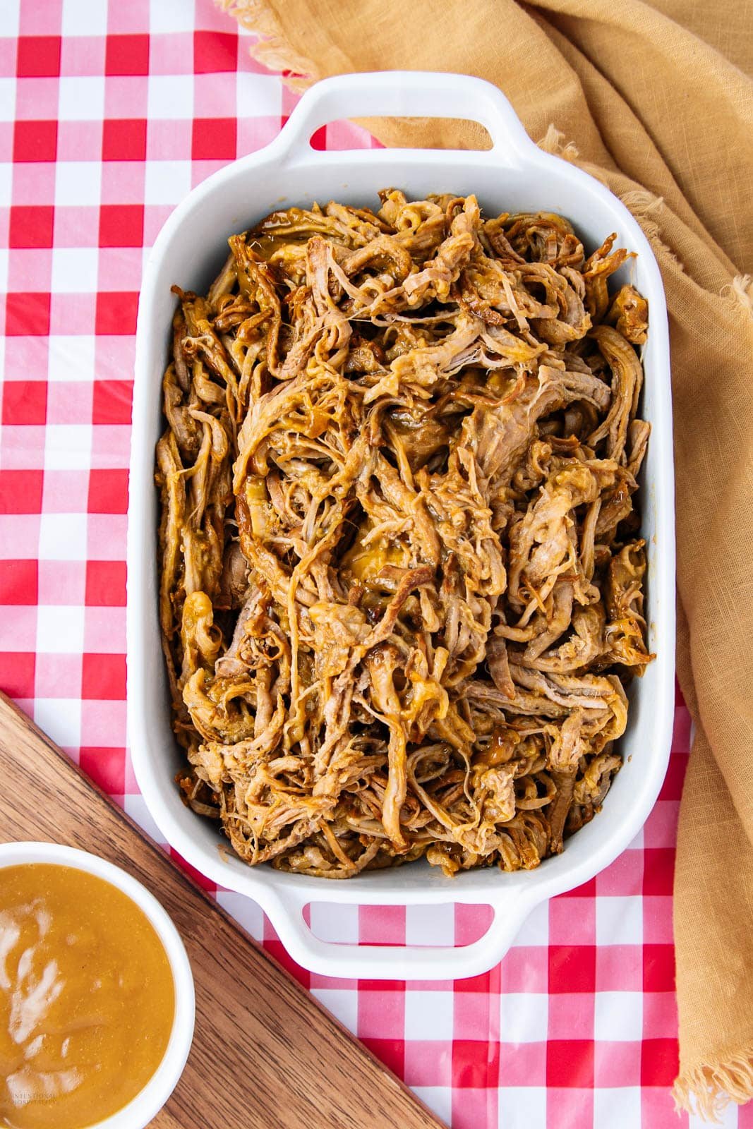 A white baking dish filled with shredded barbecue pulled pork sits on a red and white checkered tablecloth, with a bowl of sauce and a tan cloth nearby.