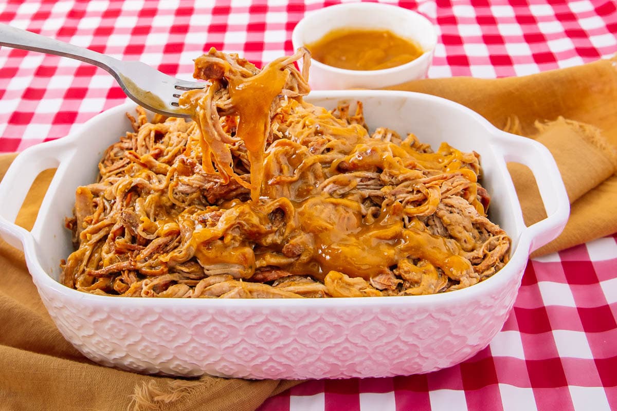 A fork lifts up saucy pulled pork from a white casserole dish, with extra barbecue sauce in a small bowl behind it. The dish sits on a red and white checkered tablecloth with a brown napkin nearby.