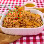 A white casserole dish filled with shredded barbecue pork, topped with golden sauce. A fork lifts a portion, dripping sauce. The dish sits on a red checkered tablecloth with extra sauce in a bowl behind it.