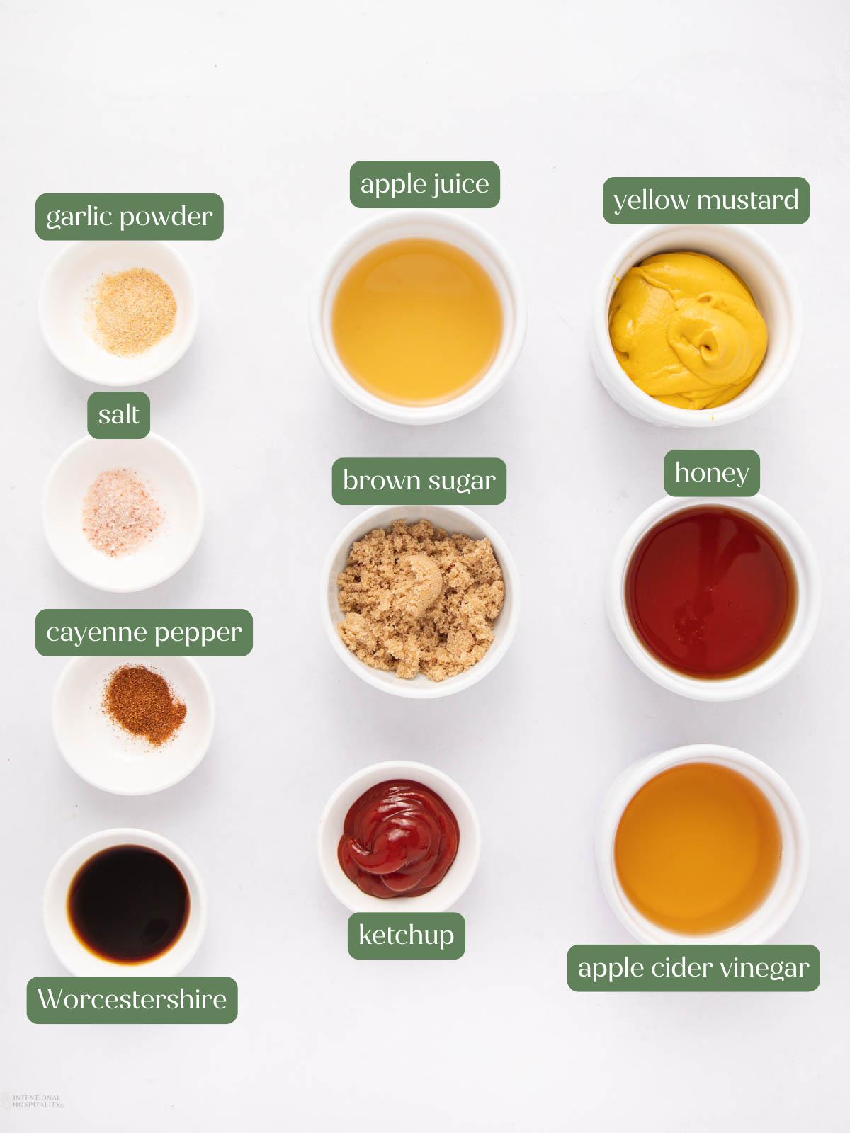 Nine small bowls arranged on a white surface, each containing a different BBQ sauce ingredient: garlic powder, apple juice, yellow mustard, salt, brown sugar, honey, cayenne pepper, ketchup, Worcestershire sauce, and apple cider vinegar.