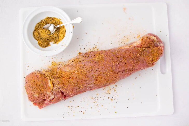 A raw pork tenderloin coated with spice rub sits on a white cutting board next to a small bowl of seasoning with a spoon.