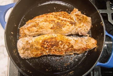 Two seasoned pork tenderloins are searing in a black cast iron skillet on a stovetop. The meat is browning and has a crust of spices, with the skillet handle visible on the left.