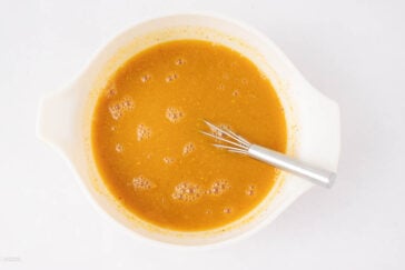 A metal whisk rests in a white mixing bowl filled with a smooth, orange-brown batter, dotted with small bubbles, against a plain white background.