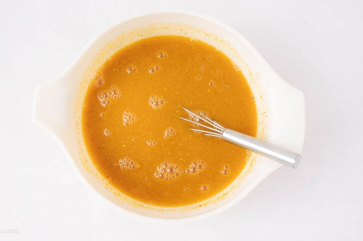 A metal whisk rests in a white mixing bowl filled with a smooth, orange-brown batter, dotted with small bubbles, against a plain white background.