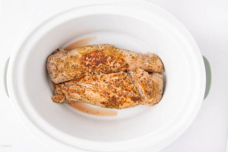 Two seasoned pork tenderloins placed side by side in a white slow cooker, ready to be cooked.