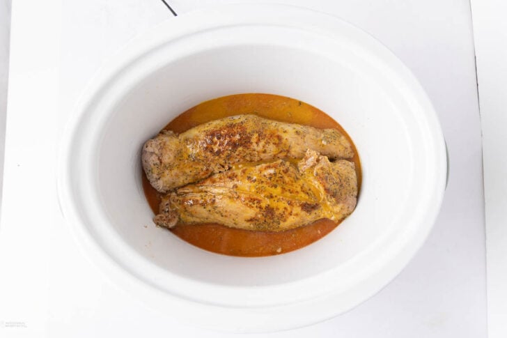 Two seasoned pork tenderloins sit in a white slow cooker, surrounded by a small amount of orange-colored sauce on the bottom.