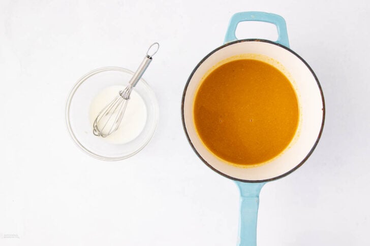 A blue-handled pot filled with orange soup sits on a white surface next to a glass bowl containing white cream and a small whisk.