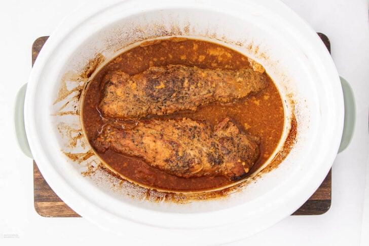 Two seasoned pork tenderloins sit in a white slow cooker, surrounded by a reddish-brown sauce. The meat appears cooked and tender, with visible spices and juices pooling around the edges.