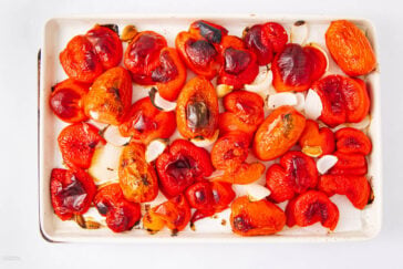 A baking sheet with roasted red bell peppers, tomatoes, onion pieces, and garlic cloves. The vegetables have charred skins and are arranged in a single layer on the tray.