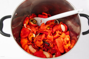 A large metal pot contains roasted red peppers and onions, with a white mixing spoon resting inside. The vegetables are partially chopped and fill most of the pot.
