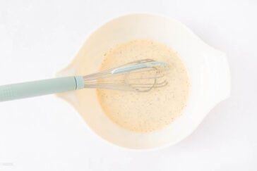 A whisk with a blue handle rests in a white mixing bowl containing a creamy, pepper-speckled sauce or batter on a white background.