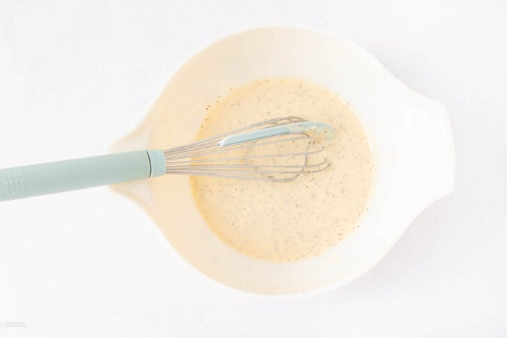 A whisk with a blue handle rests in a white mixing bowl containing a creamy, pepper-speckled sauce or batter on a white background.