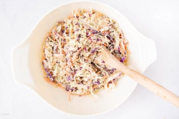 A white bowl filled with creamy coleslaw, containing shredded green and purple cabbage and carrots, with a wooden spoon resting inside on a white background.